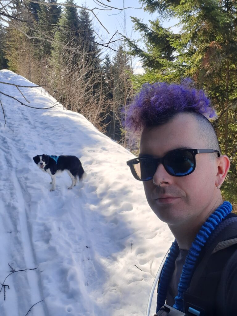 Photo d'un humain avec les cheveux violets rasés sur les cotés, des lunettes de soleil et une laisse élastique autour du cou, et un chien noir et blanc avec un harnais bleu. Le sol est couvert de neige, en arrière plan il y a des épicéas et du ciel bleu