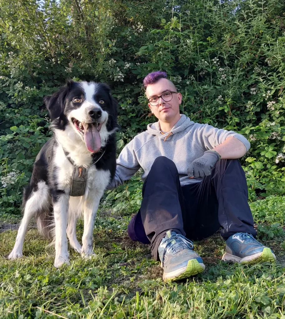 Une personne avec les cheveux violets et un chien noir et blancs assis par terre. Les 2 sont sourillant et regardent vers l'objectif